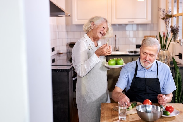 Couple de personnes âgées dans la cuisine à la maison, homme âgé en tablier est assis en train de découper des légumes frais, femme aux cheveux gris tenant des pommes dans les mains et de parler avec lui