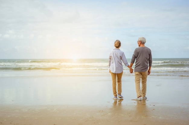 Couple de personnes âgées debout sur la plage, main dans la main au lever du soleil, plan d'assurance-vie au concept de retraite.