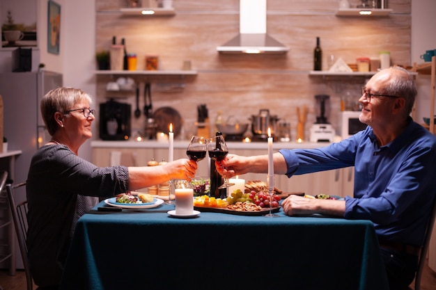 Couple de personnes âgées grillant des verres avec du vin rouge pendant le dîner. Homme et femme âgés assis à table dans la cuisine, parlant, savourant le repas, célébrant leur anniversaire dans la salle à manger.