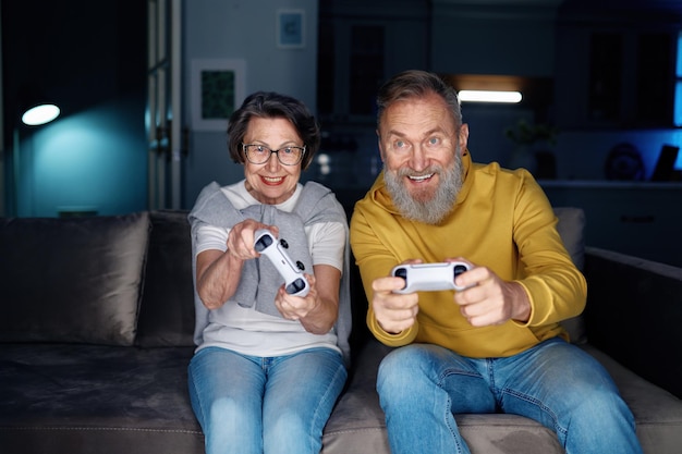 Couple de personnes âgées jouant à des jeux vidéo ensemble à la maison. Une femme et un mari âgés assis sur un canapé tenant des joysticks et se sentant excités par le temps libre le week-end