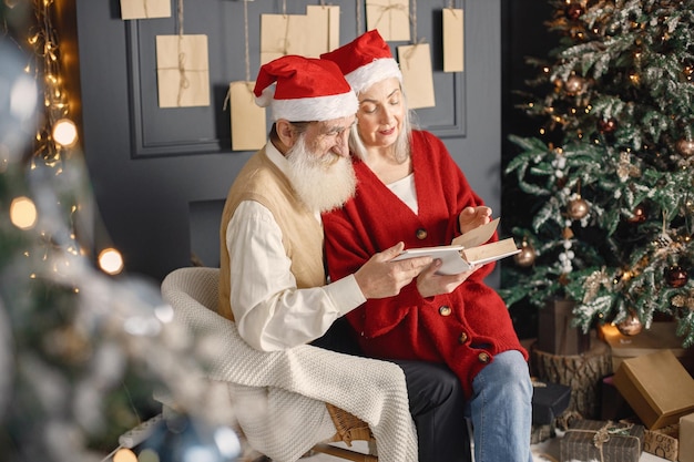 Couple de personnes âgées lisant un livre ensemble assis près de l'arbre de noël