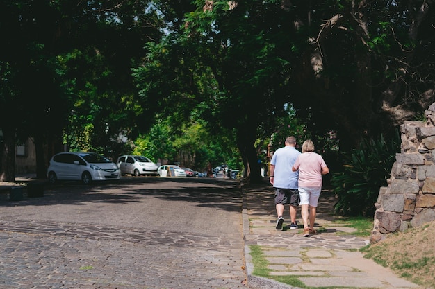 Couple de personnes âgées marchant le long du trottoir d'une rue pavée avec un tunnel d'arbres Copy space