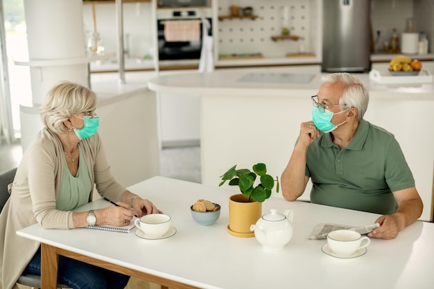 Couple de personnes âgées avec des masques faciaux parlant assis à la table à la maison