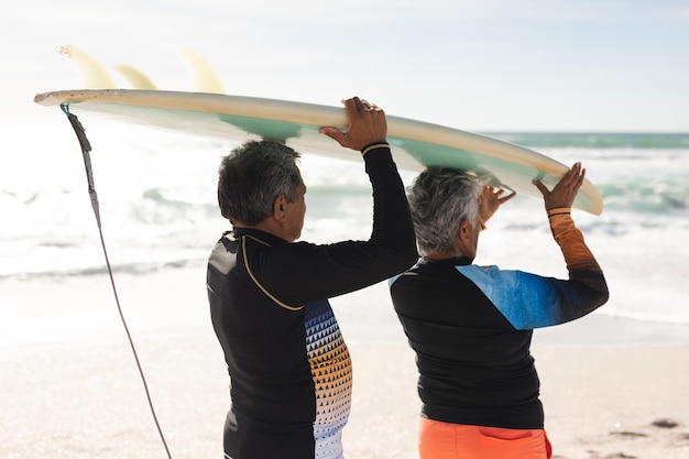 Couple de personnes âgées multiraciales marchant ensemble transportant une planche de surf au-dessus de leurs têtes sur le rivage à une plage ensoleillée