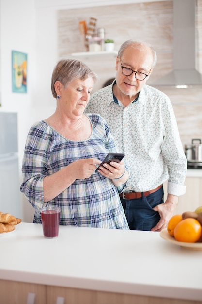 Couple de personnes âgées à la recherche sur Internet et à la recherche d'informations. Épouse et mari seniors caucasiens dans la cuisine pendant le petit-déjeuner à l'aide de la technologie des smartphones et de la connexion Internet. Femme retraitée loisirs