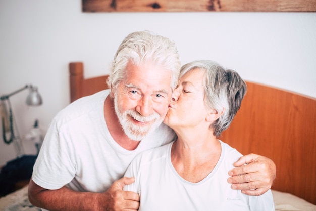 Couple de personnes âgées regardant et souriant avec amour et affect pendant qu'elle l'embrasse - adultes retraités et matures mariés dans la chambre le matin avant de se lever