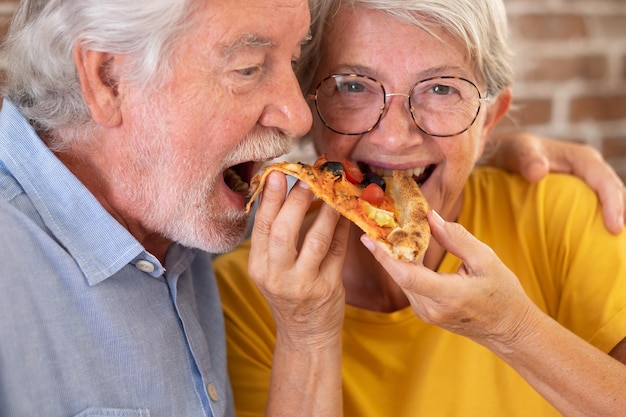 Couple de personnes âgées s'amusant ensemble en train de manger de la pizza à la maison partageant le même fond de mur de brique de tranche