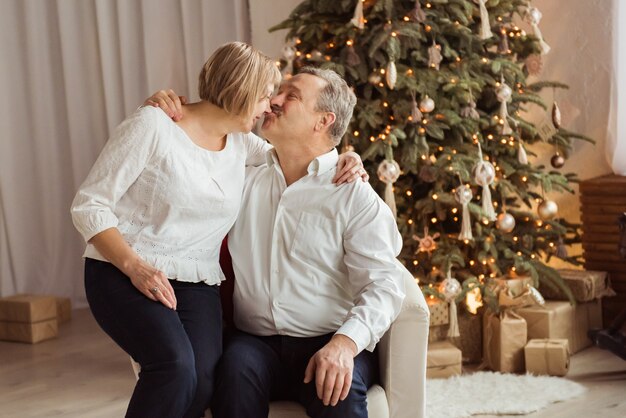 Couple de personnes âgées s'embrassant à côté de leur arbre de Noël à la maison dans le salon