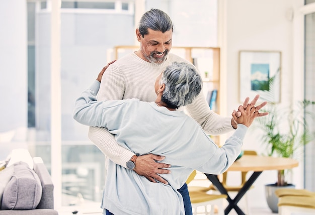 Un couple de personnes âgées s'embrassent et se tiennent la main en dansant pour l'amour, les soins ou les liens ensemble dans le salon à la maison. Un homme et une femme âgés heureux profitant d'un temps de qualité, de la retraite ou de la célébration d'un anniversaire.