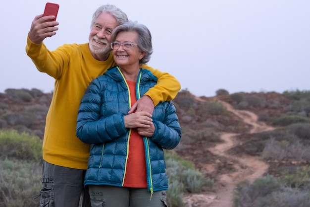 Couple de personnes âgées souriant à la campagne prenant un selfie avec un téléphone portable. Personnes âgées insouciantes habillées décontractées profitant d'une excursion dans un sentier de montagne