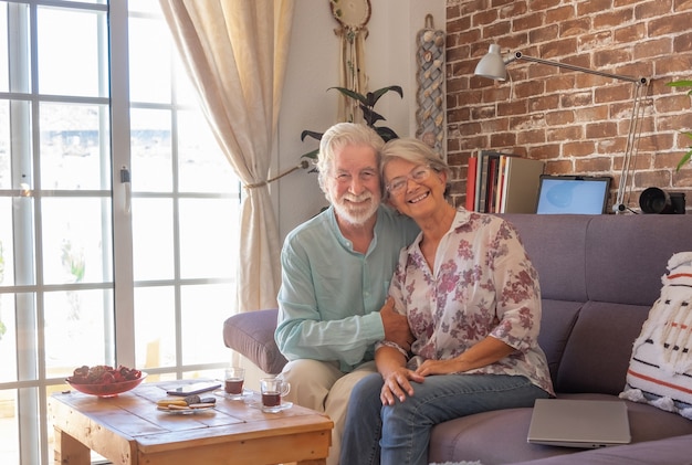 Couple de personnes âgées souriant à la maison assis sur un canapé en regardant la caméra. Mur de briques sur fond