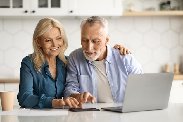 Couple de personnes âgées souriant avec ordinateur portable dans la cuisine calculant les factures à la maison