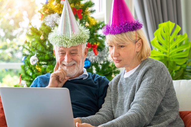 Couple de personnes âgées utilisant un ordinateur portable pour un appel vidéo pour saluer leur famille pour le festival de Noël assis sur un canapé avec décoration et arbre