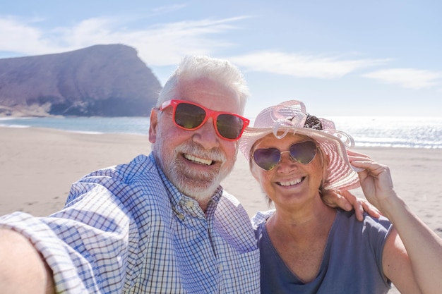 Couple de personnes âgées voyageant et prenant un selfie sur une belle plage avec une montagne en arrière-plan - souriant et appréciant de regarder la caméra avec des lunettes de soleil - mode de vie de vacances