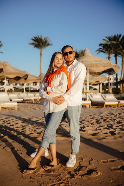 Un couple sur la plage vêtu d'une chemise blanche et d'un jean se fait un câlin et le gars porte des lunettes de soleil.