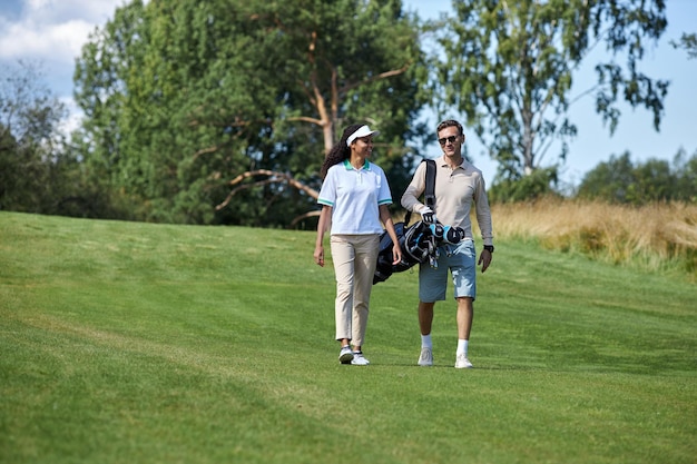 Couple portant un sac de golf marchant sur un champ vert vers la caméra et bavardant