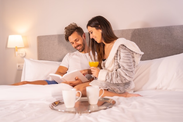 Un couple en pyjama lisant un livre au petit déjeuner de café et de jus d'orange dans le lit de l'hôtel, mode de vie d'un couple amoureux.