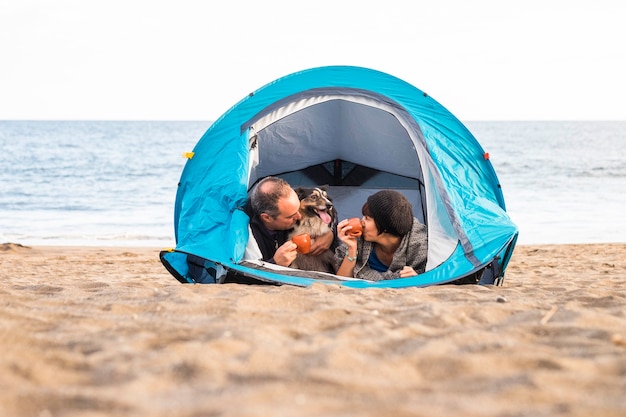 couple regardant le téléphone intelligent et s'amuser à l'intérieur d'une tente en camping gratuit sur la plage Chien border collie derrière eux en regardant la caméra. couleurs vintage et concept de famille de vacances.