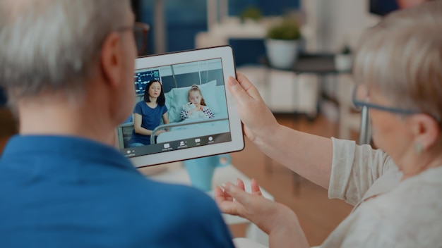 Couple à la retraite rencontrant sa famille lors d'un appel vidéo en ligne, tenant une tablette pour la communication à distance. Homme et femme plus âgés utilisant un appareil avec vidéoconférence pour parler à des proches. Fermer