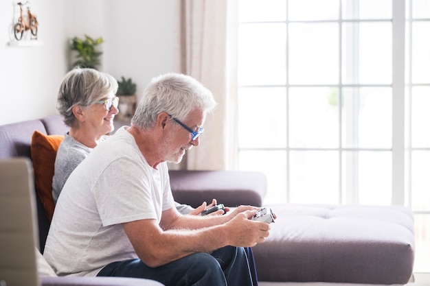 Couple de retraités caucasiens jouant à des jeux vidéo ensemble à la maison assis sur le canapé - femme mûre et homme mariés - deux se fiancent pour toujours - tenant un contrôleur ou un joystick