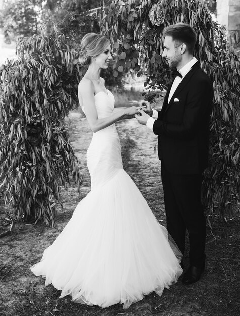 Photo un couple en robe de mariée échange des bagues dans le jardin avec une arche. mariée et le marié. photo en noir et blanc