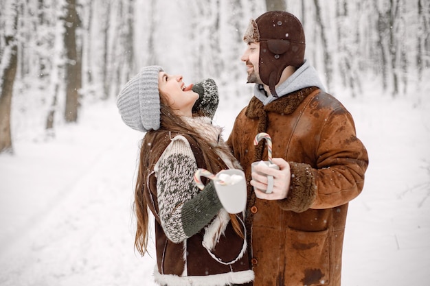 Couple romantique debout dans la forêt d'hiver étreignant et tenant deux tasses avec une boisson au cacao