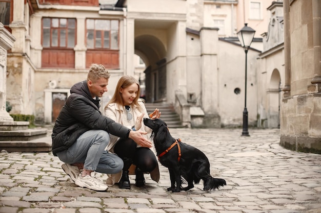 Couple romantique debout dans la rue à l'heure d'automne Homme et femme caressant un chien noir dans une rue de la vieille ville Fille portant un manteau beige et une veste noire pour homme