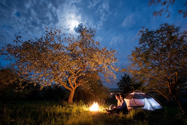 Couple romantique touristes assis devant un feu de camp près de la tente sous les arbres et le ciel nocturne avec la lune.