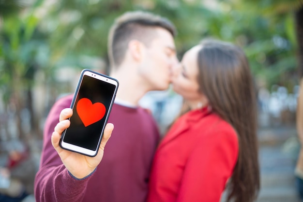 Photo couple s'embrassant tout en montrant un smartphone avec une forme de coeur dans le concept d'application de rencontres