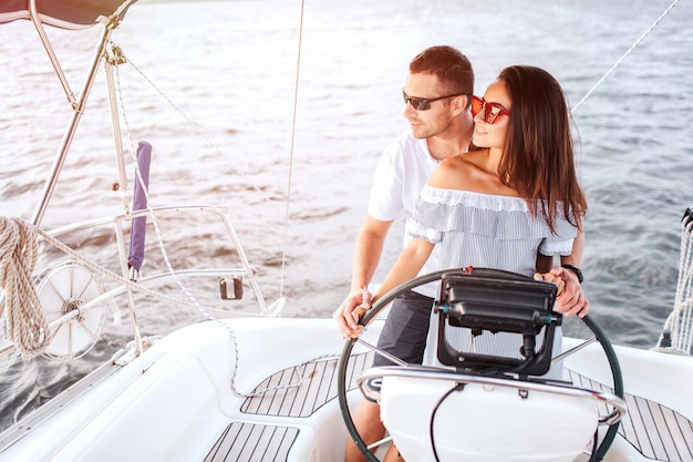 Le couple se tient ensemble et regarde à gauche. Elle garde les mains sur le gouvernail.