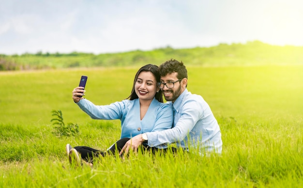 Couple souriant amoureux assis sur l'herbe prenant des selfies Jeune couple amoureux prenant un selfie sur le terrain Personnes amoureuses prenant des selfies sur le terrain avec leur smartphone