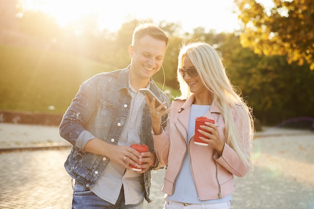 Couple souriant amoureux marchant dans le parc automne, main dans la main. Écoutez de la musique avec des écouteurs et buvez du café.