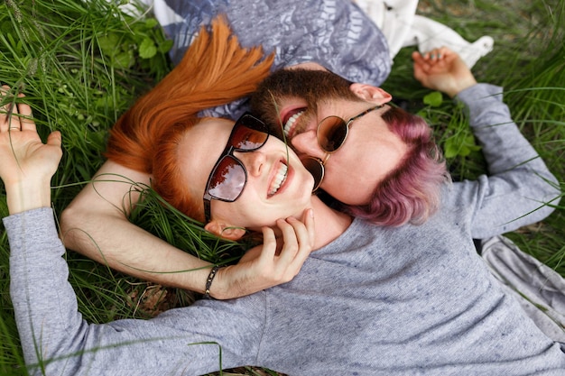 Couple souriant, assis sur une herbe verte, dans un parc, assis avec la tête tournée, dans des lunettes de soleil, regardant la caméra.