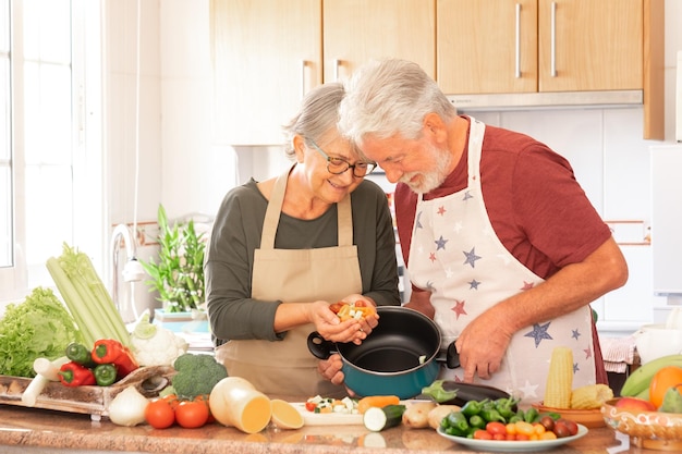 Couple souriant de personnes âgées en tabliers de chefs préparant ensemble des légumes à soupe dans la cuisine à domicile