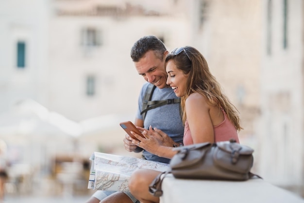 Couple souriant profitant des vacances d'été tout en étant assis sur les escaliers de la ville méditerranéenne et en explorant le plan de la ville.