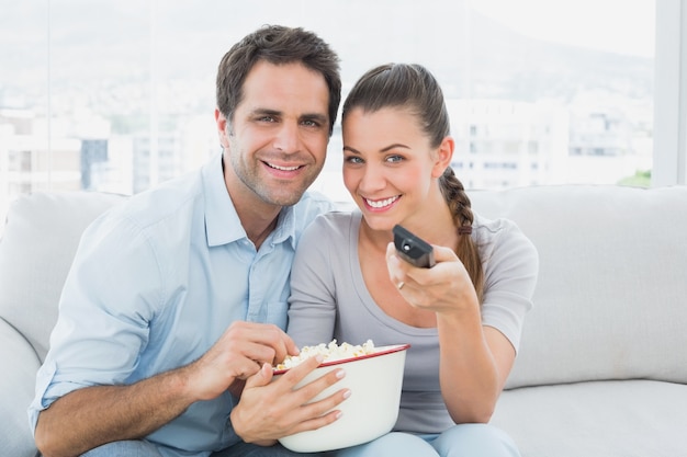 Couple souriant, regarder la télévision sur le canapé avec un bol de maïs soufflé