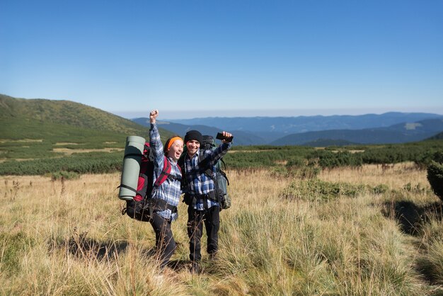 Un couple de touristes amoureux, un homme et une femme debout sur une plaine de montagne lèvent la main et crient joyeusement