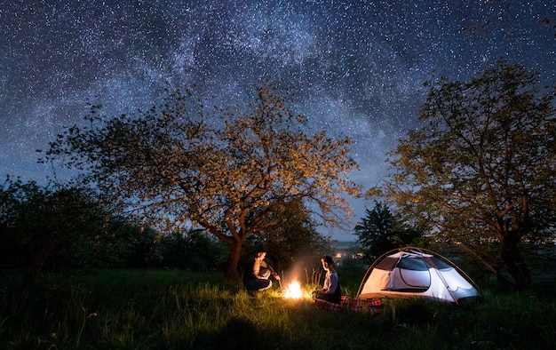 Couple de touristes assis devant un feu de camp près de la tente sous les arbres et le beau ciel nocturne plein d'étoiles et de voie lactée. Camping de nuit