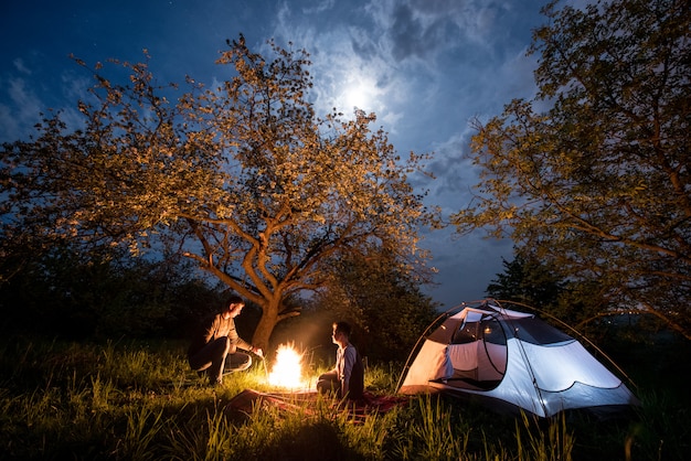 Couple de touristes assis devant un feu de camp près d'une tente sous les arbres et le ciel nocturne avec la lune. Camping de nuit