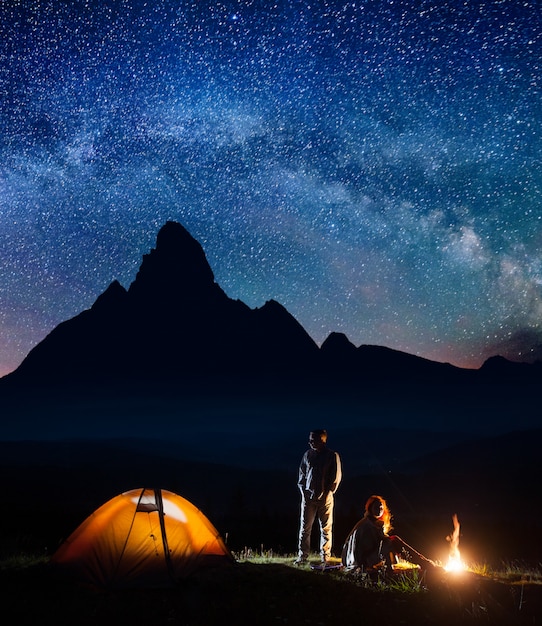 Couple de touristes romantique dans son camp la nuit près du feu de camp et tente sur fond de haute montagne et ciel étoilé