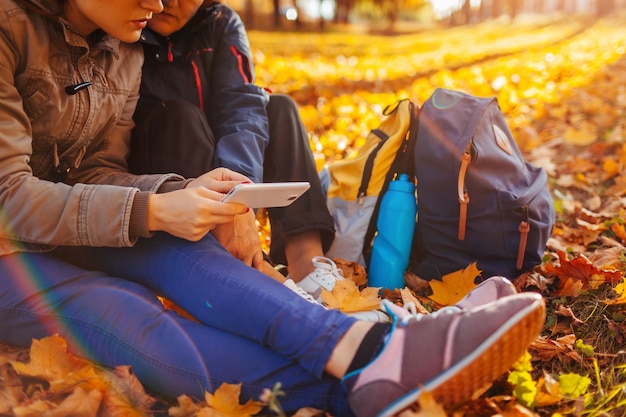 Couple de touristes avec des sacs à dos à la recherche du bon chemin à l'aide du navigateur sur téléphone dans la forêt d'automne. Les femmes se reposent