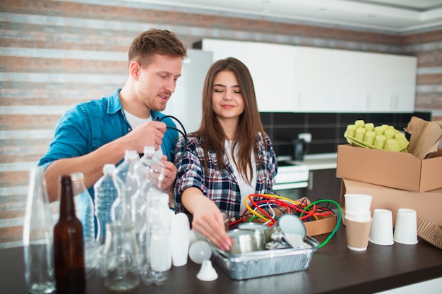 Un couple trie les ordures dans la cuisine. Les déchets doivent être envoyés pour recyclage. Il y a beaucoup de matières recyclables. Sur la table se trouvent du plastique, du verre, du fer, du papier, de vieux appareils électriques et des déchets biodégradables.