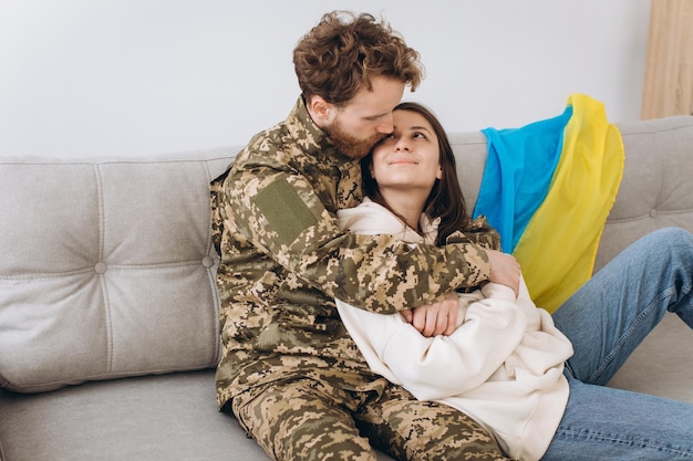 Couple ukrainien militaire en uniforme avec sa petite amie sur le canapé à la maison sur fond de drapeau jaune et bleu