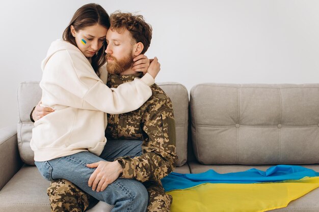 Couple ukrainien militaire en uniforme avec sa petite amie sur le canapé à la maison sur fond de drapeau jaune et bleu