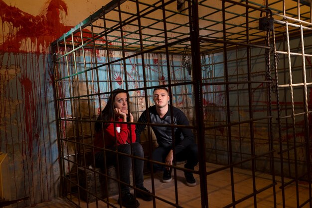 Couple de victimes d'Halloween effrayées emprisonnées dans une cage en métal avec un mur éclaboussé de sang derrière elles assises dans la terreur