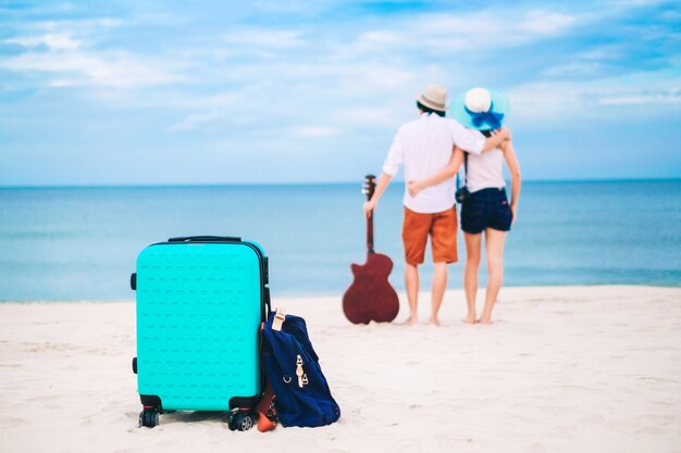 Un couple de voyageurs a un bagage et une guitare debout sur la plage.