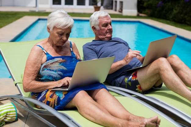 Couples aînés, utilisation, ordinateur portable, sur, chaise longue, à, piscine