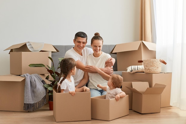 Des couples souriants positifs assis sur le sol de leur nouvel appartement et se serrant dans leurs bras, regardant des enfants dans des boîtes et s'amusant ensemble tout en emménageant dans un nouvel appartement.