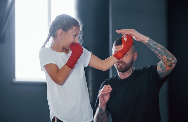 Coups de poing de base. Le jeune entraîneur de boxe tatoué enseigne la petite fille mignonne dans le gymnase.