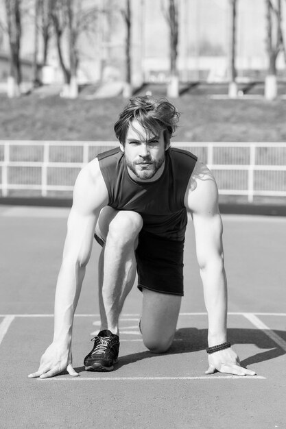 Coureur concentré athlétique barbu avec corps musclé commencer sur une piste de course ensoleillée en plein air en bleu sportswear activité d'été et sport mode de vie sain compétition défi d'entraînement